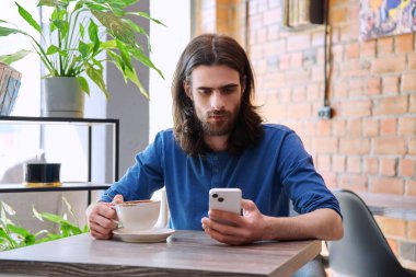 30 'lu yaşlarda şık, yakışıklı sakallı, uzun saçlı, akıllı telefon kullanan, kahve içen, kafede oturan bir adam. Mobil teknolojiler İnternet uygulamaları boş zaman iş iletişimi uygulamaları