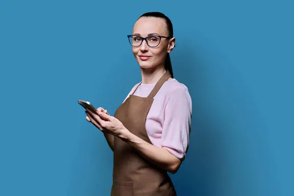 青い背景のカメラを見ているスマートフォンでエプロンの30代女性の肖像画 送信注文を受信する携帯電話のテキストを使用して笑顔の女性 テクノロジー アプリケーション サービス 中小企業 — ストック写真
