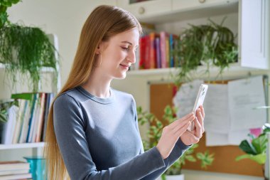 Akıllı telefon kullanan mutlu, gülümseyen genç kız, evde dikiliyor, 16,17, 18 yaşındaki kız mesajları okuyor. İletişim, eğlence, öğrenme ve alışveriş için modern dijital teknolojiler