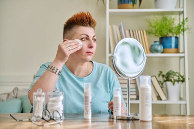 Evinde oturan aynalı orta yaşlı bir kadın. Yaşlanma önleyici cilt bakım ürünleri. Nemlendirici gıdaları temizlemek için. Güzellik çağı sağlık temizliği, 40 'lı yaşların ciltleri için tıbbi kozmetik ürünler.