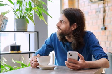 30 'lu yaşlarda şık, yakışıklı sakallı, uzun saçlı, akıllı telefon kullanan, kahve içen, kafede oturan bir adam. Mobil teknolojiler İnternet uygulamaları boş zaman iş iletişimi uygulamaları