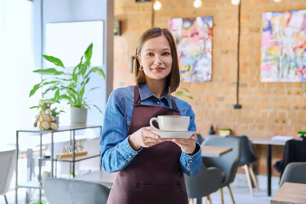 Successful young woman service worker owner in apron with cup of coffee looking at camera in restaurant cafeteria coffee pastry shop interior. Small business staff occupation entrepreneur work