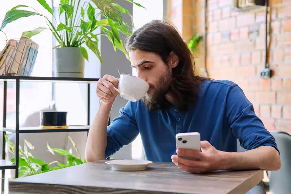 Młody Latek Przystojny Brodaty Długimi Włosami Korzystający Smartfona Pijący Kawę Zdjęcie Stockowe