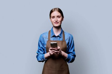 Önlüklü genç bir bayan işçi gri stüdyo arka planında akıllı telefon tutarak kameraya bakıyor. İş, iş, internet hizmetleri, mobil uygulamalar, teknoloji kavramı