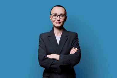 Mavi stüdyo arka planında 30 'lu yaşlarda çapraz kollu bir iş kadını portresi. Gözlüklü, takım elbiseli, kameraya bakan kendine güvenen bir kadın. İş, iş, iş, kariyer, insanlar kavramı