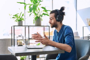 Kulaklıklı genç yakışıklı adam, kafede oturmuş laptopunu kullanarak iş görüşmesi yapıyor. Uzak sanal sanal çalışma çalışması serbest blog vlog, internet teknolojisi gençliği