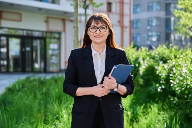 Orta yaşlı, takım elbiseli, dijital tabletli, açık hava binalı bir iş kadını. Okul eğitim merkezi yakınında poz veren kadın öğretmen, ofis çalışanı girişimci muhasebeci ekonomist bankacı finansör avukat