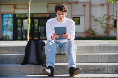 Genç yakışıklı üniversite öğrencisi dışarıda dijital tablet kullanıyor, eğitim binası geçmişi var. Eğitim, teknoloji, eğitim, 19-20 yaş gençlik kavramı