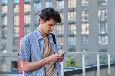 Relaxed resting handsome young male with smartphone, urban outdoor, modern building background. Student 19-20 years old using phone, texting, lifestyle, youth concept clipart