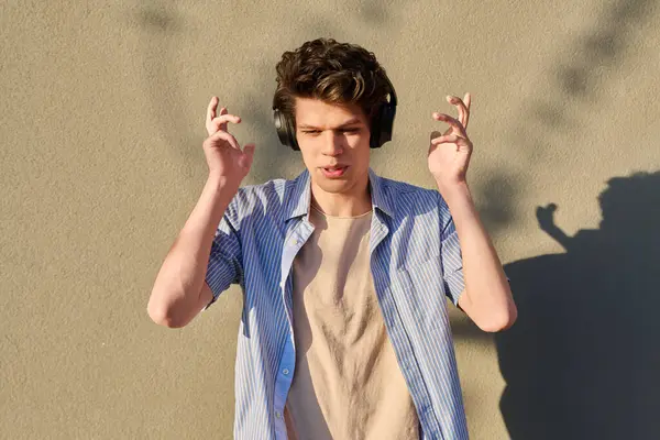 Genç yakışıklı, kablosuz kulaklıklı, açık havada güneşli gri bir duvarın arkasında müzik dinleyen bir erkek.