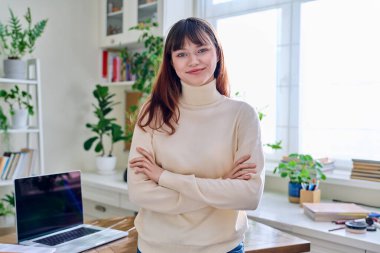 19,20 yaşındaki gülümseyen kız öğrencinin portresi. Kollarını kavuşturmuş, kameraya bakıyor, evin içinde. Gençlik, yaşam tarzı, üniversite konsepti