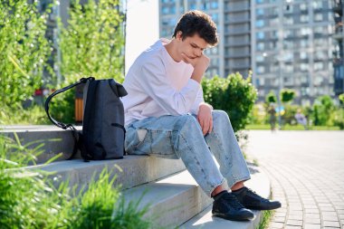 Üzgün, mutsuz, genç bir erkek merdivenlerde oturuyor. Sırt çantasıyla oturan, başı elle tutulan üniversite öğrencisi. Sorunlar, zorluklar, depresyon, gençlerin zihinsel sağlığı