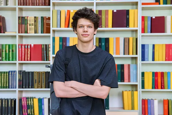 Portrait of confident handsome college student guy with crossed arms inside library of educational building. Education, youth, lifestyle concept