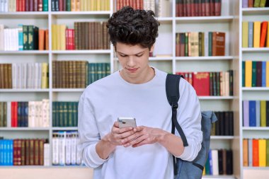 Sırt çantalı genç bir erkek öğrenci. Akıllı telefon kullanıyor. Üniversite binasında, kütüphanede. Teknolojiler, mobil eğitim uygulamaları uygulamaları, hizmetler, e-öğrenim kavramı