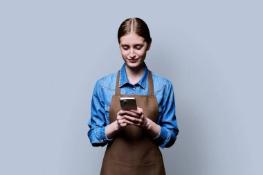 Gri stüdyo arka planında akıllı telefon kullanan önlüklü genç bir bayan işçi. İş, iş, internet hizmetleri, mobil uygulamalar, teknoloji kavramı