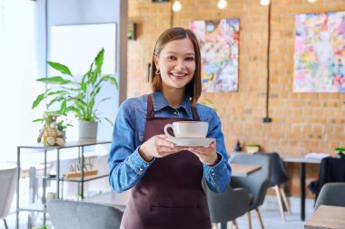 Önlüklü başarılı genç bayan servis çalışanı bir fincan kahveyle kafeteryanın içindeki kahvecinin kamerasına bakıyor. Küçük işletme işgali girişimciliği