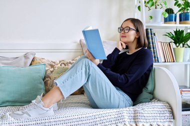 Kitap okuyan genç bir kadın, evdeki kanepede oturuyor, kendini kurguya kaptırmış. Edebiyat hobisi, eğitim, yaşam tarzı, gençlik kavramı