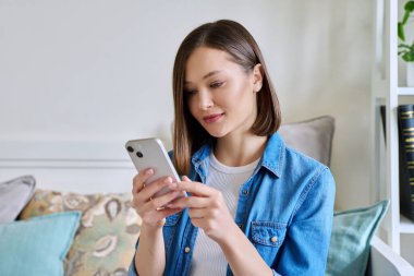 Evdeki koltukta oturan akıllı telefonu kullanan genç bir kadın. Kadın mesajlaşma görüntüsü, mobil uygulamalar teknoloji iş eğitimi için internet hizmetleri eğlence iletişim alışveriş yaşam tarzı