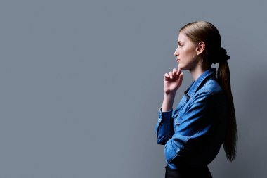 Profil görüntüsü, özgüveni yüksek genç bir kadın gri stüdyo arkaplanı arıyor, reklam metni için kopyalama alanı. Kot gömlekli, güzel sarışın, moda tarzında.