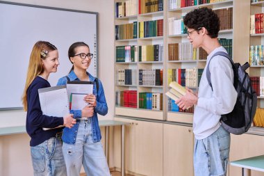 Bir grup genç lise öğrencisi kütüphane sınıfında. 16-17 yaşlarındaki üç okul çocuğu birlikte gülüyor. Eğitim, ergenlik, arkadaşlık iletişimi konsepti