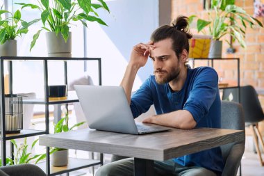 Genç, ciddi bir adam laptopla çalışıyor ve birlikte çalıştığı kafede oturuyor. 30 'ların yakışıklı sakallı adamı bilgisayar ekranına bakıyor. Boş zamanların teknolojisi iş eğitimi eğitimleri iletişim insanları