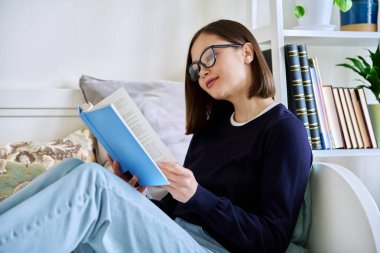 Kitap okuyan genç bir kadın, evdeki kanepede oturuyor, kendini kurguya kaptırmış. Edebiyat hobisi, eğitim, yaşam tarzı, gençlik kavramı