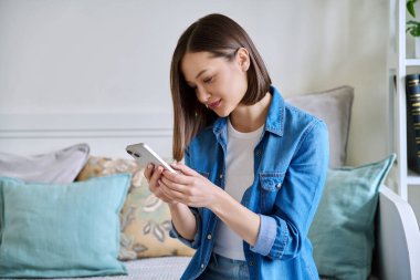 Evdeki koltukta oturan akıllı telefonu kullanan genç bir kadın. Kadın mesajlaşma görüntüsü, mobil uygulamalar teknoloji iş eğitimi için internet hizmetleri eğlence iletişim alışveriş yaşam tarzı