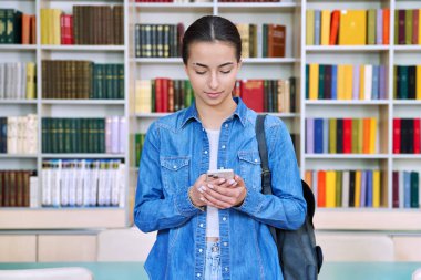Sırt çantalı genç kız öğrenci akıllı telefon kullanıyor, lise binasında, kütüphanede. Teknolojiler, mobil eğitim uygulamaları uygulamaları, hizmetler, e-öğrenim kavramı