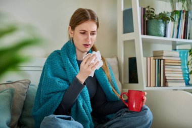 Genç hasta kadın sıcak battaniyenin altında oturup öksürüyor burnunu mendile sümkürerek öksürüyor, evdeki kanepede oturuyor. Soğuk algınlığı, sonbahar kışı mevsimsel solunum hastalıkları, tedavi ve bakım