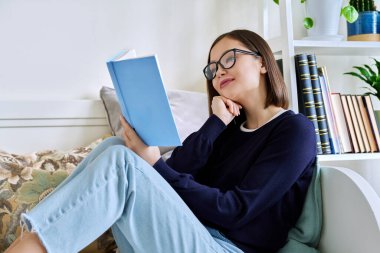 Kitap okuyan genç bir kadın, evdeki kanepede oturuyor, kendini kurguya kaptırmış. Edebiyat hobisi, eğitim, yaşam tarzı, gençlik kavramı