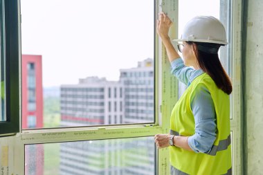 Koruyucu kask giyen kadın endüstriyel işçiler pencereden yeni binalara bakıyor. İnşaat mühendisliği, emlak satışları ofis daireleri, teknik meslekler.