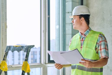 Güvenlik kasklı genç sanayi işçisinin portresi. İnşaatla ilgili mühendislik belgeleriyle birlikte çalışıyor. İnşaat onarım servisi geliştirme çalışma konsepti