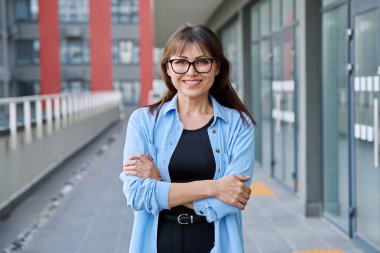 Açık havada gülümseyen, kendine güvenen olgun bir kadının şehir arka planında çekilmiş bir portresi. Yaşam tarzı, orta yaş, iş çalışma konsepti