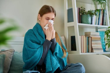 Genç hasta kadın sıcak battaniyenin altında oturup öksürüyor burnunu mendile sümkürerek öksürüyor, evdeki kanepede oturuyor. Soğuk algınlığı, sonbahar kışı mevsimsel solunum hastalıkları, tedavi ve bakım