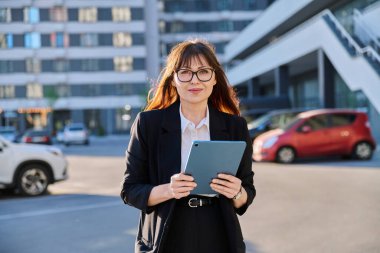 Siyah takım elbiseli dijital tabletli başarılı iş kadını modern şehrin arka planında, açık havada kameraya bakıyor. İş, girişimcilik sigorta satış reklamları danışmanlığı