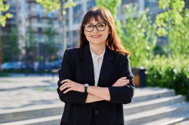 Orta yaşlı, kendine güvenen, takım elbiseli, çapraz kollu, şehir tarzında poz veren bir kadının portresi. Ofis çalışanı iş kadını öğretmen girişimci ekonomist bankacı finansör avukat