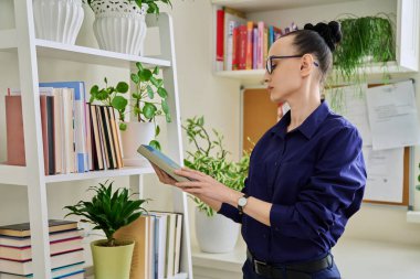 Evde, kitaplığın yanında oturan orta yaşlı bir kadın öğrenim öğretmek için kitap seçiyor. Yaşam tarzı, edebiyat, iş kitapları, boş zaman, insan kavramı