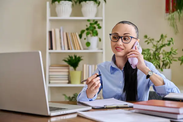 stock image Business woman talking on mobile phone sitting at desk with laptop computer. Serious confident 30s female working with business papers. Work, workplace, people concept