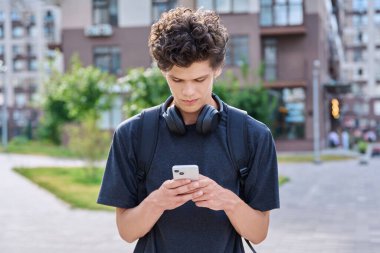 Modern şehir sokaklarında akıllı telefon kullanan genç bir adam. 20 yaşındaki yakışıklı erkek öğrenci telefon ekranına bakıyor. Gençlik, teknoloji mobil uygulamaları, internet iletişim hizmetleri, yaşam tarzı