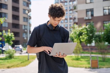 Sırt çantası kulaklıklı genç bir üniversite öğrencisi. Laptop kullanıyor. Açık şehir geçmişi var. Yakışıklı, gülümseyen, 20 yaşında, dizüstü bilgisayarında kıvırcık saçlı.