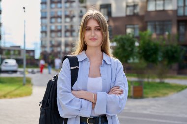 Sırt çantasıyla kameraya bakan sarışın genç kızın açık hava portresi. Kollarını çapraz yapmış kendine güvenen güzel bir kız, şehir tarzı. Gençlik, yaşam tarzı, eğitim lisesi