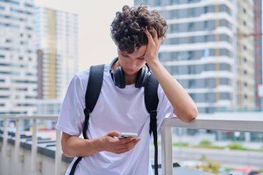 Üzgün, mutsuz, genç bir adam akıllı telefondan haber mesajı okuyor. Modern gökdelenler. Kolej öğrencisi elleriyle başını tutuyor. Sorunlar, depresyon, gençlerin akıl sağlığı