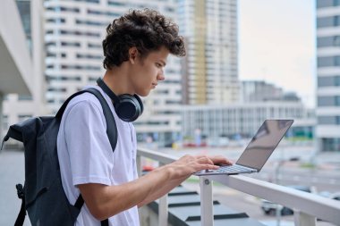 Genç erkek, dizüstü bilgisayar kullanıyor, modern şehir gökdelenleri geçmişi var. Yakışıklı bir öğrenci, 19-20 yaşlarında, boş zamanlarında bilgisayar kullanıyor, okuyor, çalışıyor. Teknoloji, gençlik, eğitim, kentsel tarz kavramı