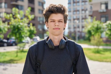 Dışarıdaki kameraya bakan yakışıklı genç adam, şehir arka planı. 20 yaşında, siyah tişörtlü, kıvırcık kulaklıklı, mutlu bir öğrenci. Gençlik, yaşam tarzı, moda tarzı, eğitim