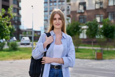 Sırt çantasıyla kameraya bakan sarışın genç kızın açık hava portresi. Kollarını çapraz yapmış kendine güvenen güzel bir kız, şehir tarzı. Gençlik, yaşam tarzı, eğitim lisesi