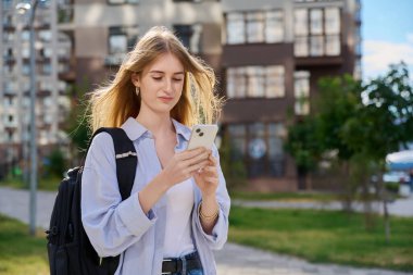 Akıllı telefon kullanan, modern şehir geçmişi olan genç sarışın bir kadın. Ergen öğrenci gezmek için mobil uygulama uygulamaları kullanarak mesajlaşıyor. Teknoloji İnternet Eğitim Gençlik Usulü