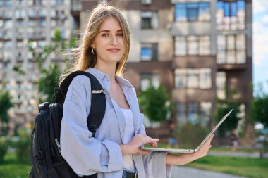 Genç, çekici bir üniversite öğrencisi dışarıda dizüstü bilgisayar kullanıyor, modern şehir geçmişi var. Eğitim, teknoloji, eğitim, 17-18 yaş gençlik kavramı