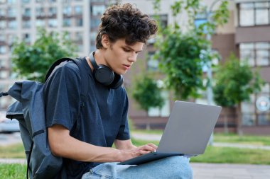 Şehirde dizüstü bilgisayar kullanan genç bir erkek. Yakışıklı bir öğrenci, 19-20 yaşlarında, boş zamanlarında bilgisayar kullanıyor, okuyor, çalışıyor. Teknoloji, gençlik, eğitim, kentsel tarz kavramı