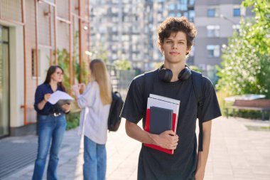Kendinden emin, gülümseyen üniversite öğrencisinin portresi, sırt çantalı, ders kitaplı, dışarıda eğitim binasının yakınındaki kameraya bakan genç bir erkek. Eğitim, eğitim, 19.20 yaş gençlik kavramı