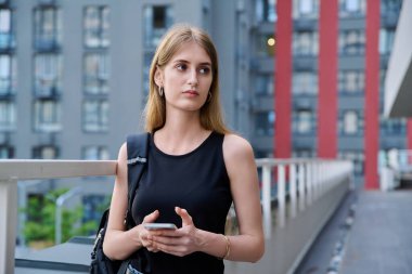 Akıllı telefon kullanan genç sarışın kadın, modern şehir geçmişi. Ergen öğrenci gezmek için mobil uygulama uygulamaları kullanarak mesajlaşıyor. Teknoloji İnternet Eğitim Gençlik Usulü